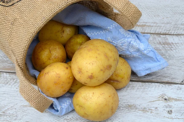 Biologische verse ongekookt aardappelen in een jute zak op een oude verweerde houten rekken van witte achtergrond met vintage blauwe keuken theedoek met lege kopie ruimte — Stockfoto