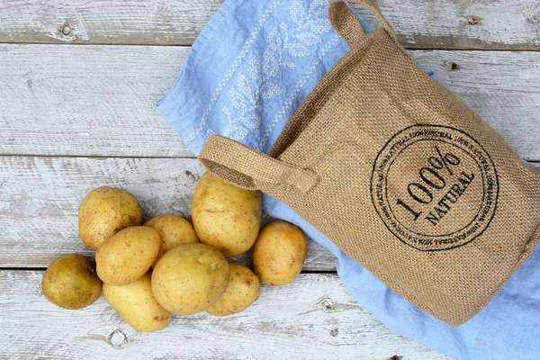 Biologische verse ongekookt aardappelen in een jute zak op een oude verweerde houten rekken van witte achtergrond met vintage blauwe keuken theedoek met lege kopie ruimte — Stockfoto