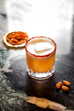 Buzlu bir bardakta portakal kokteyli, büyük bir buz küpü, paprika kabuğu kenarlı, bademle servis edilir.