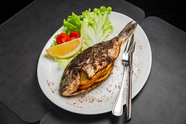Portakalla doldurulmuş ızgara dorado balığı, marul, limon ve kirazlı domates, bıçak ve çatalla süslenmiş.