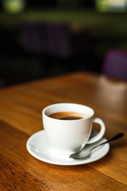 Beyaz bir fincan kahve, beyaz çay tabağı, kaşık, ahşap masa, bir kafede.