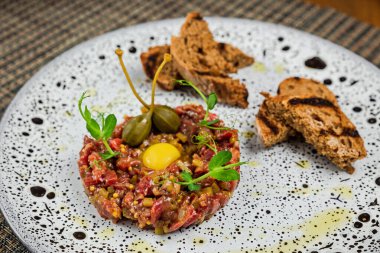 Sığır bifteği tartar, bıldırcın yumurtası sarısı ve kapari, ızgara kızarmış ekmek ile servis edilir, yakın plan, beyaz bir tabakta.