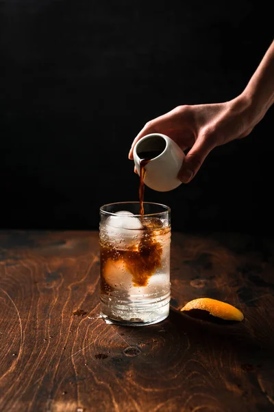 オレンジジュースでエスプレッソ強壮剤を準備します 小さな白い缶からエスプレッソを注ぐ手は氷球と強壮剤で満たされたハイボールガラスに — ストック写真