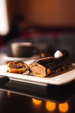 Beyaz bir tabakta çikolatalı ekler, tatlı bıçağı, çatal ve bir fincan kahvenin sade dikey fotoğrafı. Yansımalı karanlık masa. Seçici odak.