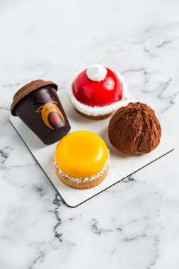 Açık beyaz mermer zemin üzerinde bir krema seti. Sarı mango tartelette, kırmızı beyaz tartelette, iki çikolatalı tatlı.