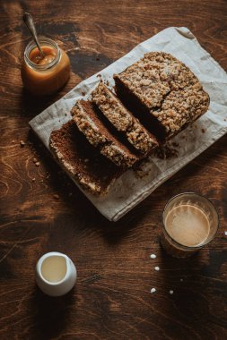 Tahta kesim tahtasında çikolatalı muz ekmeği ve keten peçete, kremalı bir bardak kahve, bir sürahi süt ve bir kavanoz tuzlu karamel. Tahta masa.. 