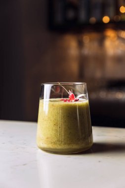 A glass of cold kiwi and banana smoothie cocktail garnished with an edible flower, marble table.