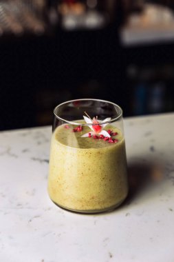 A glass of cold kiwi and banana smoothie cocktail garnished with an edible flower, marble table.