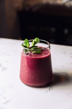 A glass of cold forest fruit berry smoothie cocktail garnished with a mint sprig, marble table.