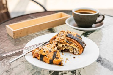 Bir fındık ve çikolatalı kruvasan yakın plan, beyaz tabakta ikiye bölünmüş, bir fincan sade kahve, bir çatal bıçak seti, bir kafe terasındaki cam masada.