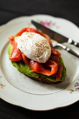 Somonlu ve haşlanmış yumurtalı ekmekli avokado tostu. Ahşap zemin, gümüş çatal bıçak takımı. Sığ alan derinliği, doğal ışık.