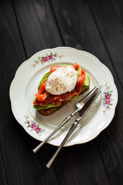 Somonlu ve haşlanmış yumurtalı ekmekli avokado tostu. Ahşap zemin, gümüş çatal bıçak takımı. Sığ alan derinliği, doğal ışık.