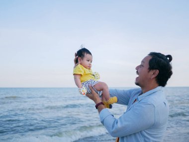 Mutlu Asyalı aile tatilleri, baba yazın sahilde şirin bir bebek tutuyor, aile deniz gezisi.