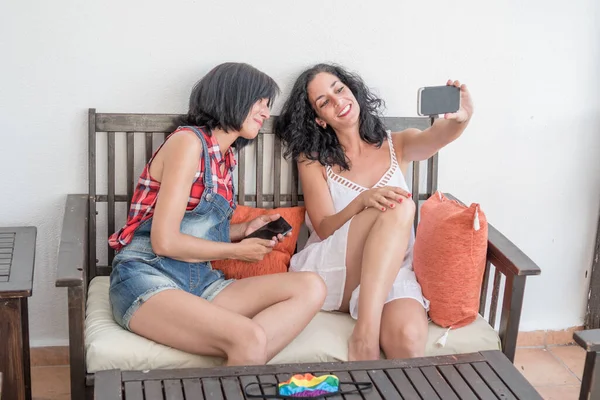 two young women laugh and have fun while taking a selfie with their mobile sitting in the chill out of the terrace of the house