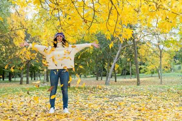 young woman with curly black hair in white sweater and knit hat has fun throwing dry leaves that have fallen from the trees in a park in autumn