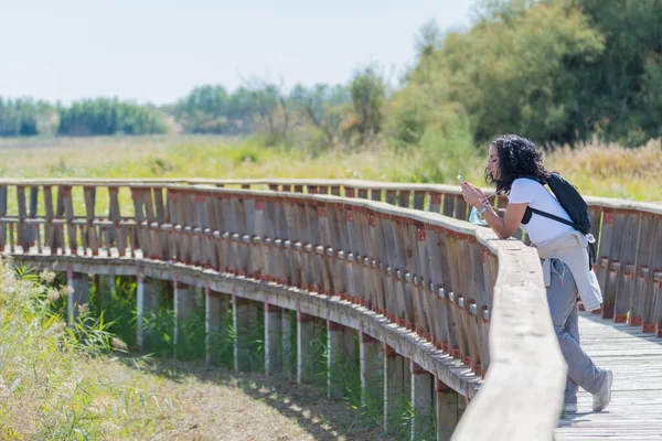 young hiker woman with curly black hair looking at her cellphone while taking a walk on a wooden bridge on a sunny day