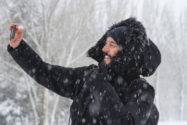 Sakallı hippi adam kendini soğuktan korumak için cep telefonu ve şapkası ile selfie çekerken kışın arka planda karlı ağaçlarla dolu bir parkta kar yağıyor.