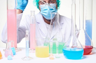 Laboratuvar teknisyeni eldiven, maske ve laboratuvar önlüğü giyiyor. Laboratuvarda deney tüpleri ve maddeler üzerinde çalışıyor.