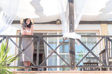  Kıvırcık saçlı ve şapkalı güzel esmer kadın yaz aylarında güneş ışınlarıyla aydınlatılan dağ evinin balkonundan sabahları tefekkür eder.