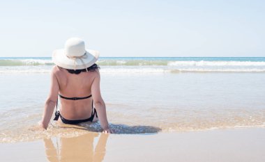 Şapkalı ve siyah bikinili genç bir kadın sahilde oturup ufka bakıyor.