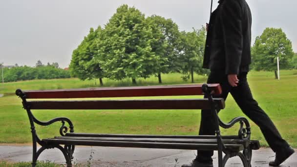 El hombre se acerca al banco y se sienta mientras llueve . — Vídeos de Stock