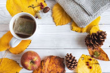 Sonbahar mevsimi kompozisyonu sarı yapraklar, kozalaklar, elma, bir kupa sıcak çay, fındık, kırmızı böğürtlen ve beyaz ahşap arka plan üzerine süveter.