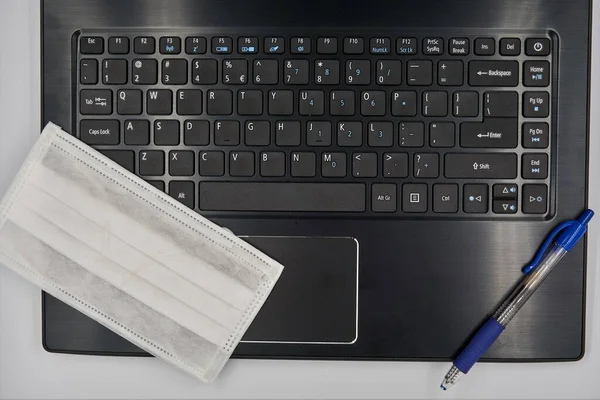 Laptopun üstüne beyaz cerrahi maske ve mavi kalem.