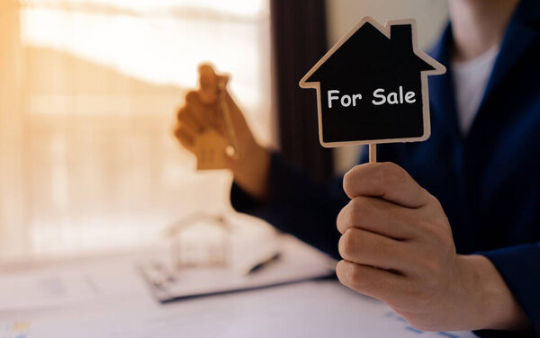 Home Seller or Realtor real estate holding key and showing house label with the text "For Sale". Background sunlight.
