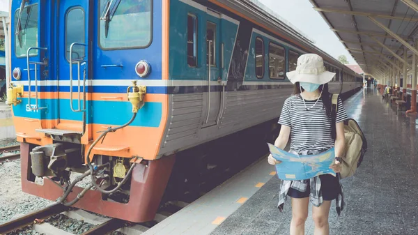 Maskeli genç bir bayan turist tren istasyonuna doğru yürüyor ve seyahat bilgileri için bir turist haritasına bakıyor. Yeni Normal seyahat ve turizm kavramı.