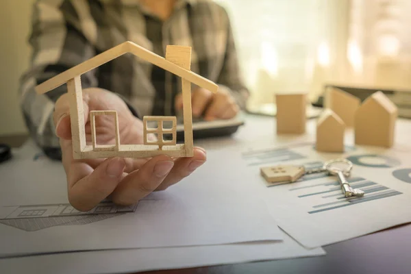 Banka memuru ya da emlakçılar, arka planda ev tasarımları dokümanı, hesap makinesi ve anahtarla birlikte maket ev modelini masada gösteriyorlar..