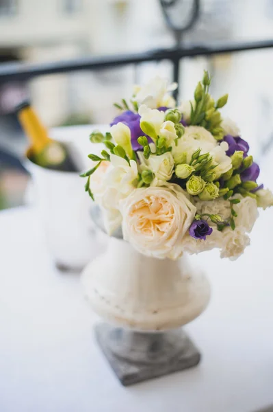 View of bridal bouquet with white and green roses, violet flowers and green leaves in decorative vase and champagne on blurred background