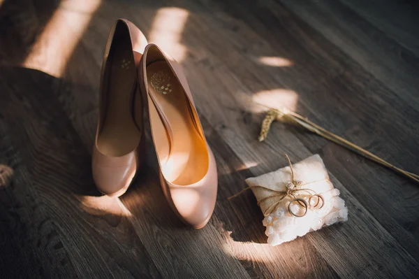 Бежевые свадебные туфли, лежащие на деревянном полу с пшеничными ушами и подушкой с обручальными кольцами сбоку — стоковое фото