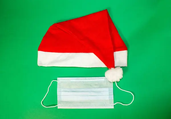 緑の背景には 医療用マスクとサンタクロースの帽子 クリスマス休暇中の保護 上からの眺め — ストック写真