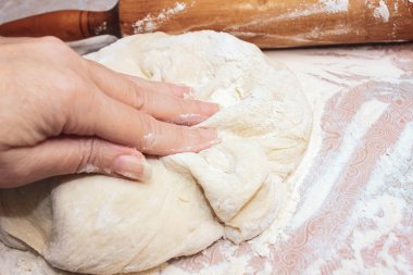 Bir kadının eli hamur mayasına basar, un içinde masanın üzerinde, merdanenin yanında. Yakın çekim, seçici çekim.