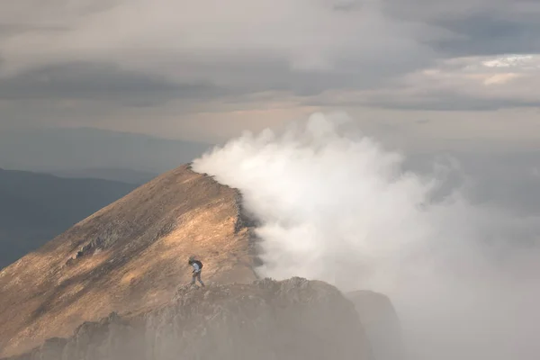 Epic view of mountain hiker standing on the top of the cliff and distant mountain ridge lighten by soft sunlight from one side and covered by thick fog from another