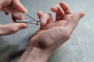Close-up of a person using a metal nail clipper to trim fingernails, focus on hands and grooming tool on a gray textured surface. High quality photo