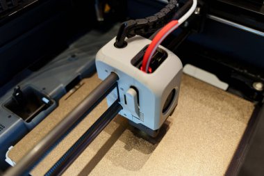 A close-up view of a 3D printer in action, showing the print head and rails, with wires attached, inside the machine's enclosure. High quality photo