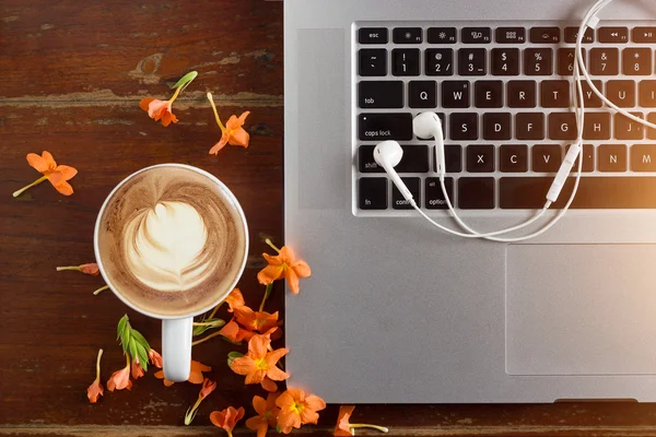 Dizüstü bilgisayar, kulaklık ve bir fincan kahve latte eski ahşap masa üzerinde çiçeklerle — Stok fotoğraf