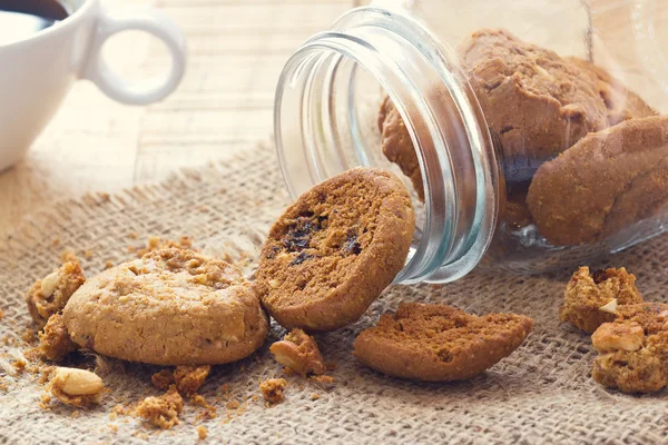Шоколадное печенье в стеклянной банке на мешке и кофе на деревянном столе — стоковое фото
