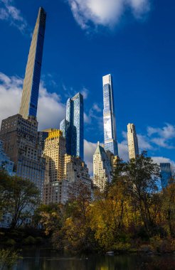 Central Park Gölü, seyahat, turizm, gayrimenkul, finans ve NYC markaları için ideal, parlak mavi gökyüzü altında altın sonbahar ağaçları tarafından çerçevelenmiş şık Manhattan gökdelenlerini yansıtıyor