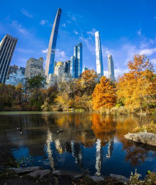 Central Park Gölü, seyahat, turizm, gayrimenkul, finans ve NYC markaları için ideal, parlak mavi gökyüzü altında altın sonbahar ağaçları tarafından çerçevelenmiş şık Manhattan gökdelenlerini yansıtıyor