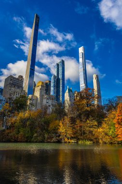Central Park Gölü, seyahat, turizm, gayrimenkul, finans ve NYC markaları için ideal, parlak mavi gökyüzü altında altın sonbahar ağaçları tarafından çerçevelenmiş şık Manhattan gökdelenlerini yansıtıyor