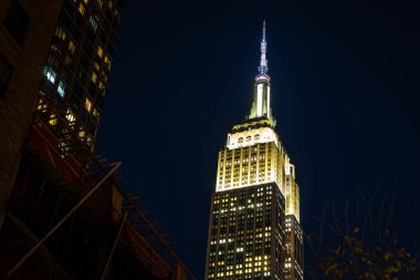 Aydınlatılmış Empire State Binası, klasik cephelerle ve parlayan ışıklarla çerçevelenmiş Manhattan Caddesi üzerinde gece yükselir; seyahat, turizm, şehir markalaşması ve emlak reklamları için ideal