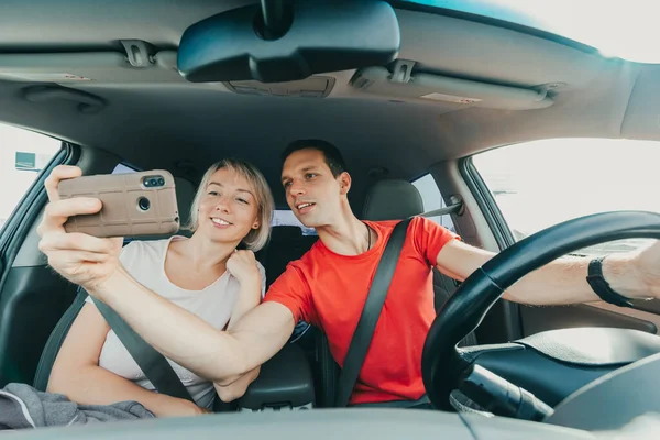 Pareja feliz en el amor en viaje hace selfie usando teléfono inteligente y coche de conducción. Gente sonriente dentro de un vehículo nuevo usando cinturones de seguridad —  Fotos de Stock