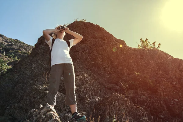 Sırt çantalı genç bir kadın, dağlardaki kayalarda dururken dürbünle bakıyor. Seyahat ve macera konsepti