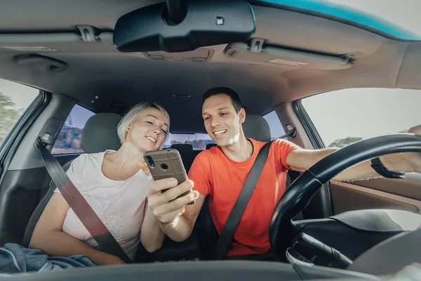 Jong gelukkig stel rijden auto en het gebruik van de smartphone tijdens het rijden op de weg. Man en vrouw samen op zoek naar mobiele telefoon — Stockfoto