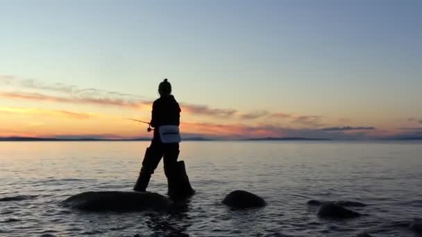 Γυναίκα ψαράς ρίχνει δόλωμα με περιστρεφόμενη ράβδο στο νερό για να πιάσει πέστροφα από την ακτή κατά τη διάρκεια υπέροχο ήσυχο ηλιοβασίλεμα στη λίμνη γλυκού νερού — Αρχείο Βίντεο