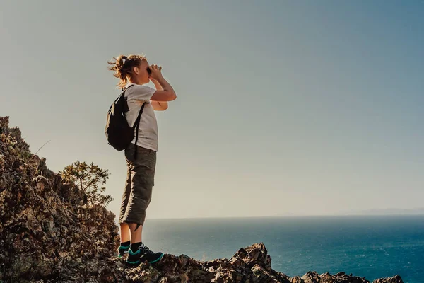 Escursionista giovane ragazza con lo zaino alla ricerca in binocolo godendo di una vista spettacolare dalla cima della montagna sull'infinito del mare, cielo blu — Foto Stock