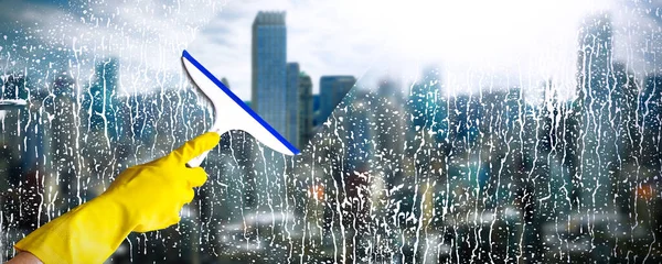 Cleaning windows with skyscraper on background. Hand in yellow glove hold cleaning squeegee. Cleaning service concept. — Stock Photo, Image