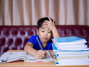 Ödev yapan ve yanında bir yığın kitapla ahşap bir masada kitap okuyan çocuk kırmızı bir kanepe ve krem perdedir..